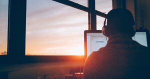 Persona di spalle davanti al monitor di un computer accanto a una finestra al tramonto