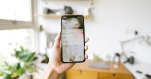 Mano che tiene uno smartphone acceso in una stanza luminosa con scrivania e piante sullo sfondo