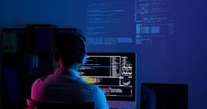 Sviluppatore visto di spalle davanti a due monitor con codice e finestre di terminale in un ufficio illuminato di blu